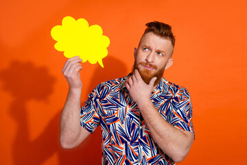 Portrait of clever man wear print shirt thoughtfully look empty sapce hold yellow bubble hand on chin isolated on orange color background