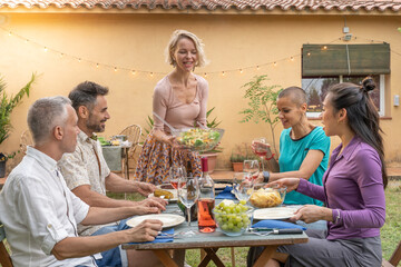 Multiethnic friends laughing with big smile around the table. Woman serves the food at house patio....