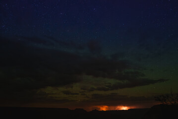 Night sky with stars over dark landscape, silhouettes of mesas against lighter sky. Clouds and...