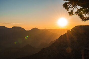 Sunset landscape with rugged mountains fading into distance, warm golden glow in the sky. Silhouetted tree branch frames the scene, lens flares add interest. Peaceful, majestic mood.