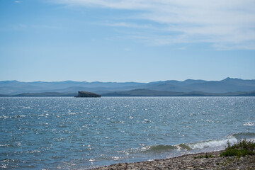 Water waves landscape, view of Lake Baikal and island in distance, piece of land with grass on rocks, on horizon faintly visible mountains of opposite shore under beautiful sky, sunny summer day.