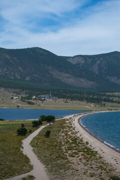 Cape Uyuga on Lake Baikal. Spit, part of land, protrudes deep into blue water surface on sides. Side top view. Waves hitting rocky shore, trees growing on the shore, forested mountains in background