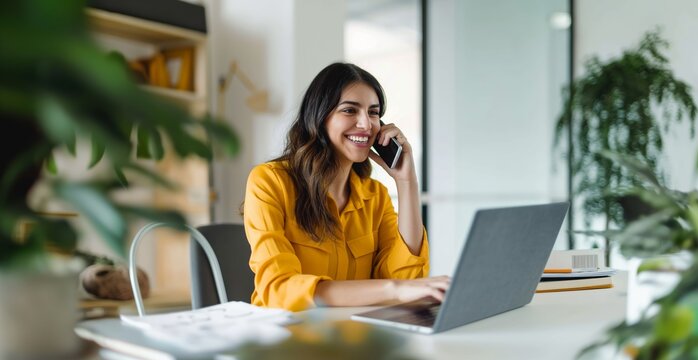 A woman is talking on her cell phone while sitting at a desk with a laptop. She is smiling and she is enjoying her conversation,wearing yellow clothes