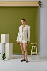 A stylish young woman wearing a white blazer and shorts stands confidently in a modern studio with green backdrop. Her look is completed with a chic necklace and black shoes.