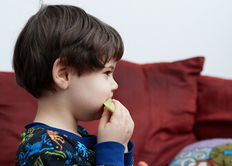 cute baby boy eating a cucumber on the couch in his dinasaur pajamas