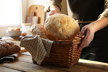 Man holding wicker basket with different types of bread at wooden table indoors, closeup