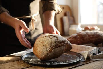 Man holding loaf of fresh bread at wooden table indoors, closeup