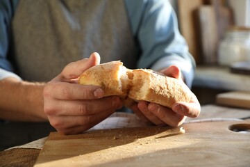 Man breaking loaf of fresh bread at wooden table indoors, closeup