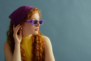 Fashionable redhead woman wearing purple headscarf, trendy sunglasses, yellow blouse,silver earrings, posing on blue background. Close up studio fashion portrait. Copy, empty, blank space for text
