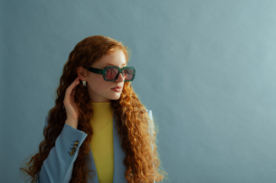Fashionable redhead confident woman wearing trendy green sunglasses, silver earrings, yellow blouse, blazer, posing on blue background. Studio fashion portrait. Copy, empty, blank space for text - Powered by Adobe