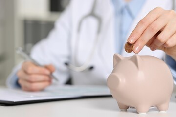 Doctor putting coin into piggy bank while making notes at white table indoors, closeup
