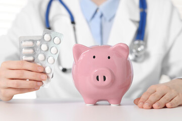 Doctor with pills and piggy bank at white table, closeup