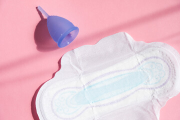 Blue menstrual cup lies next to sanitary pad on vibrant pink background, illustrating modern approach to feminine hygiene.
