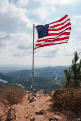 American flag on flagpole waving against hilly backdrop. Rugged terrain leads to flagpole, cloudy...