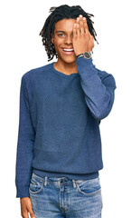 Fototapeta premium Young african american man wearing casual winter sweater covering one eye with hand, confident smile on face and surprise emotion.