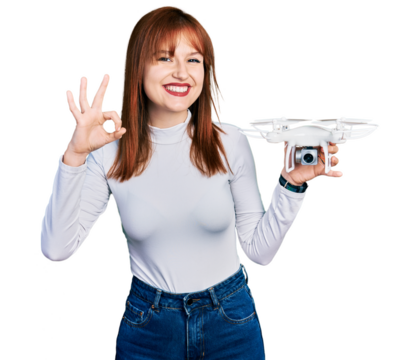 Redhead young woman holding drone doing ok sign with fingers, smiling friendly gesturing excellent symbol