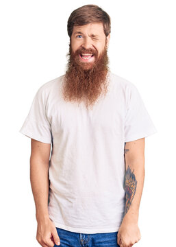 Handsome young red head man with long beard wearing casual white tshirt winking looking at the camera with sexy expression, cheerful and happy face.