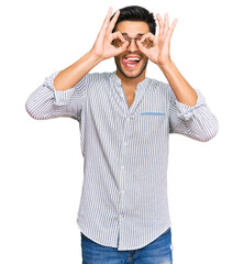 Young handsome man wearing casual clothes and glasses doing ok gesture like binoculars sticking tongue out, eyes looking through fingers. crazy expression.