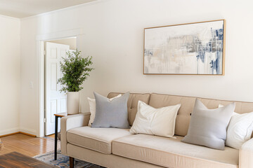 A light and airy living room with a beige sofa, grey pillows, and an abstract painting on the wall above it. The walls have white paint and there is wooden flooring in the space.