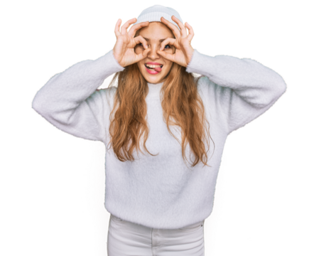 Young caucasian girl wearing wool sweater and winter cap doing ok gesture like binoculars sticking tongue out, eyes looking through fingers. crazy expression.