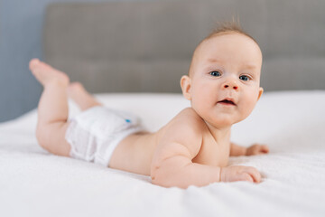 Portrait of cute healthy newborn baby lying in bed trying to crawl waving hand at home. Front view of adorable little kid having good mood. Rising girl, first steps in crawling after hand plank.