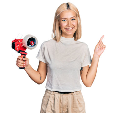 Beautiful young blonde woman holding packing tape smiling happy pointing with hand and finger to the side