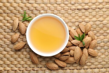 Almond oil in bowl, nuts and leaves on wicker table, flat lay