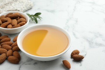 Almond oil in bowl and nuts on white marble table, closeup. Space for text