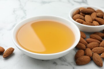 Almond oil in bowl and nuts on white marble table, closeup