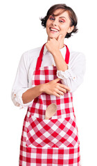 Beautiful young woman with short hair wearing professional cook apron looking confident at the camera with smile with crossed arms and hand raised on chin. thinking positive.