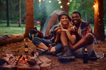 Happy couple eating hot dogs while camping in nature and looking at camera.