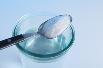 Glass of water and spoon with baking soda on light blue background, closeup