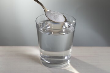 Spoon with baking soda over glass of water on table against light grey background, closeup