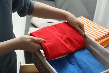 Woman putting folded clothes into storage basket indoors, closeup