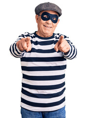 Senior handsome man wearing burglar mask and t-shirt pointing to you and the camera with fingers, smiling positive and cheerful