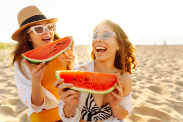 Female friends on vacation at the beach in summer eating a watermelon with the sea in the background, sitting on the sand at sunset. People, lifestyle, travel, nature and vacations concept.