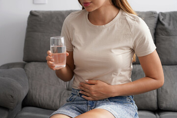 Costiveness. Unreconizable young woman suffering from abdominal pain holds a glass of water in her hand at home. - obrazy, fototapety, plakaty