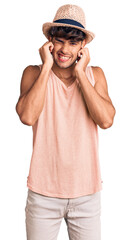 Young hispanic man wearing summer hat covering ears with fingers with annoyed expression for the noise of loud music. deaf concept.
