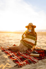 Young woman sits at picnic on the beach drinks a hot drink from a thermos. A girl enjoying beautiful view of the sea. Travel, weekend, relax and lifestyle concept
