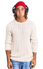 Young hispanic man listening to music using headphones puffing cheeks with funny face. mouth inflated with air, crazy expression.