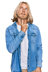 Caucasian man with blond long hair wearing casual denim jacket thinking concentrated about doubt with finger on chin and looking up wondering