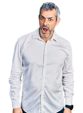 Middle age hispanic with grey hair wearing casual white shirt in shock face, looking skeptical and sarcastic, surprised with open mouth