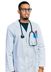 Young african american man wearing doctor uniform and stethoscope in shock face, looking skeptical and sarcastic, surprised with open mouth