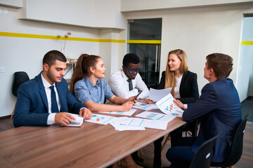 Multicultural team led by female boss analyzes graphs in office boardroom. Group engages in discussion at corporate meeting. Mixed-race professionals collaborate on project strategy.