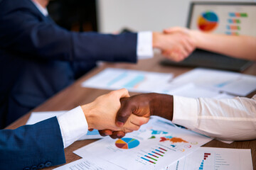 Multiracial professionals handshake closing at office meeting. Charts on table, laptop in...