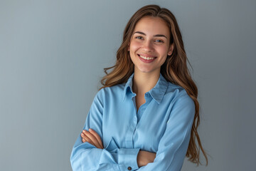 With a smile on her face, a confident young businesswoman in a blue shirt looks into the camera, presenting her professionalism and charisma, standing with her arms crossed against a gray background.