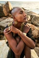Non-binary black person in luxury dress on rocks in ocean . Trans ethnic fashion model wearing...