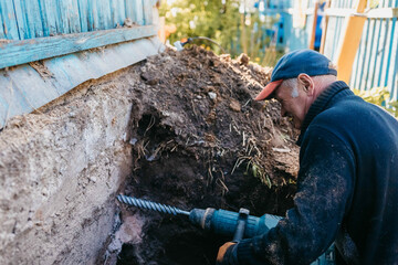 male worker drills the foundation with a hammer drill for sewerage in a country house.