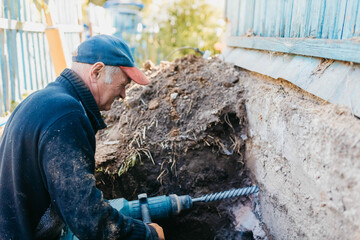 male worker drills the foundation with a hammer drill for sewerage in a country house.