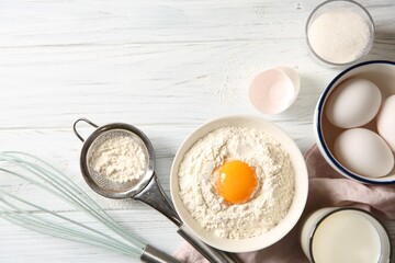Flour with yolk in bowl and other ingredients for dough on white wooden table, flat lay